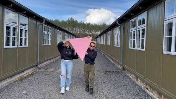 Lotus Lemaire and George Saxon holding up a huge pink triangle, situatet between two barracks in the memorial.